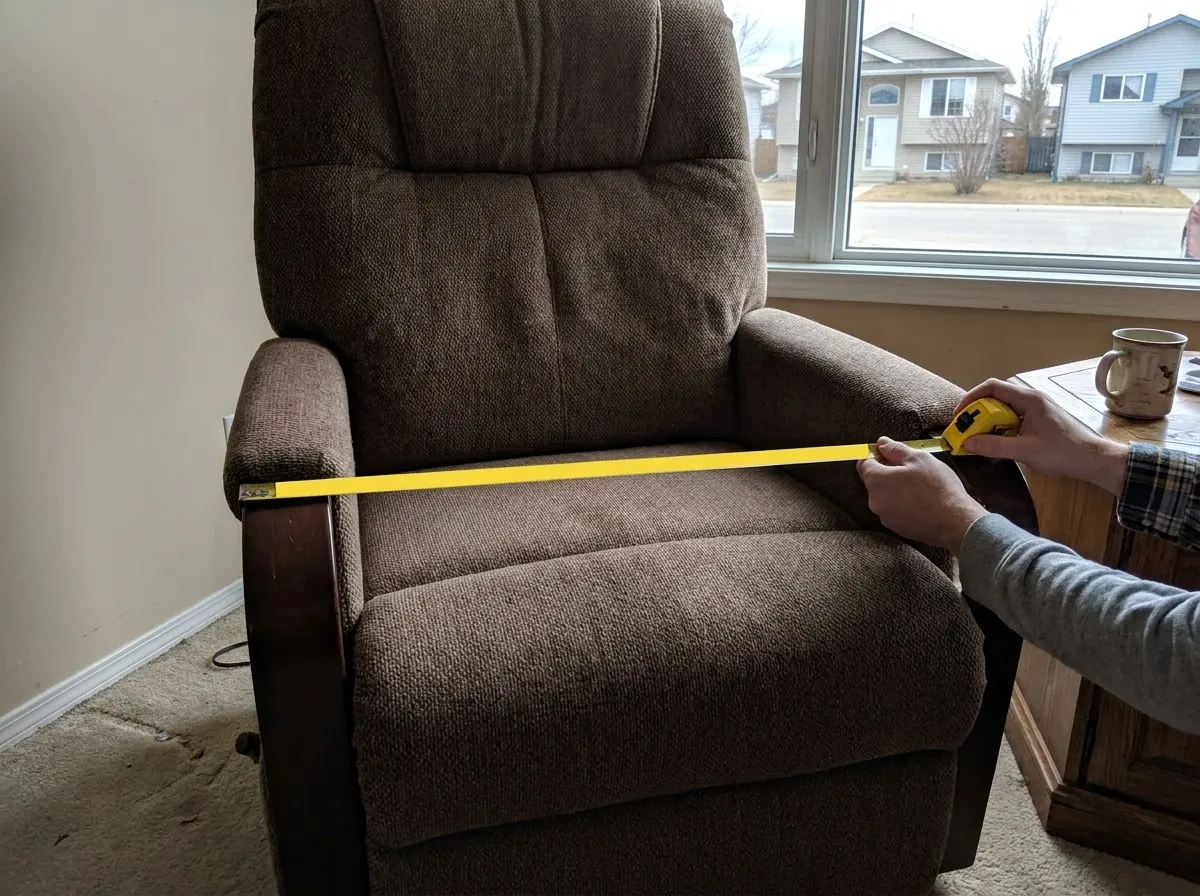 Hands stretching a tape measure across the seat of a wide upholstered lift chair between the armrests.