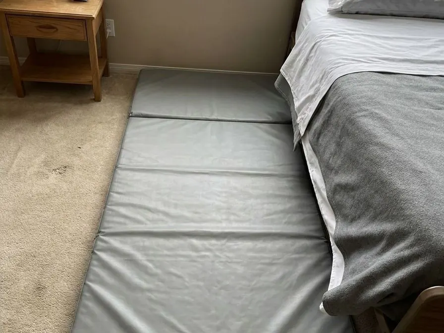 Looking down at a thick grey padded fall mat lying on bedroom carpet along the edge of a made hospital bed.